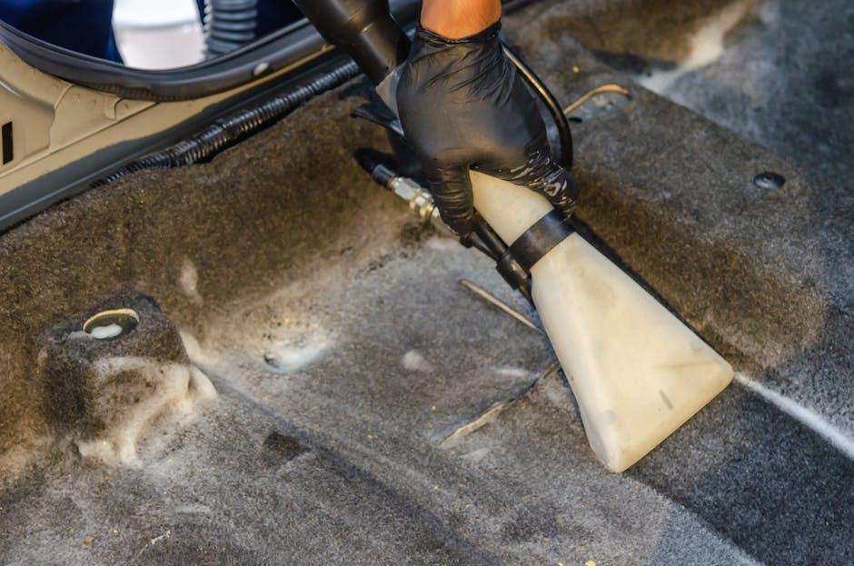 A person using a long-handled mop to clean a gray carpeted floor in a darkened indoor space, possibly a conference room or auditorium, with a large white projection screen overhead. The individual is